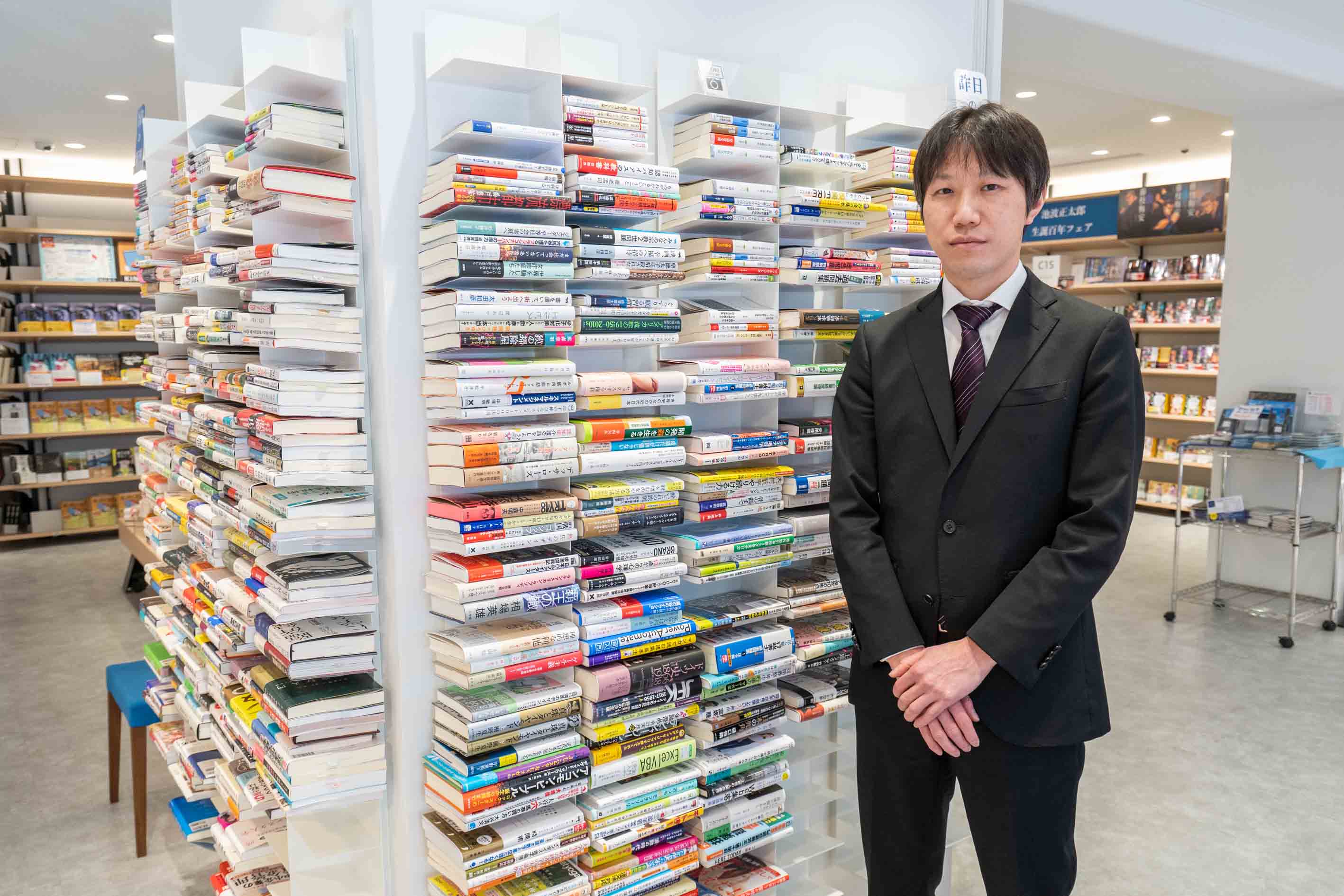 紀伊國屋書店新宿本店の吉野裕司副店長。新刊を縦に並べずに横に積み上げる「BOOK WALL」の前で