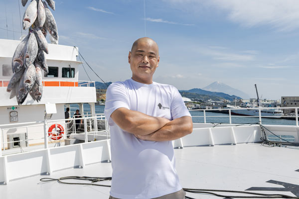 クレーンでつり上げたマグロを背に立つ小山涼太氏（写真＝小林 淳）