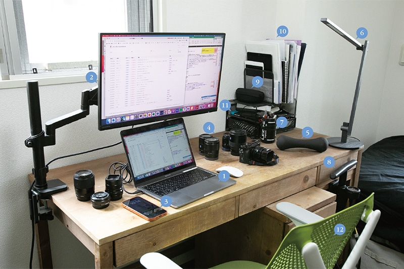 2年半前に横浜ワールドポーターズ内の家具店で購入した木製デスクを勉強机に。「いろいろ置きたいので奥行きのあるタイプを選びました」。1仕事用のMacBook Pro。「社員の大半はWindowsを使っていますが、僕はApple好きなのでMacに」。(2)I-O DATAのモニター。「モニターの下のデスクスペースを使えるようにアーム式に」。(3)公私で使うiPhone。(4)集中力が切れたときにいじるカメラとレンズ。(5)体がこり固まったときに持ち上げる3kgのダンベル。(6)アマゾンで購入したLEDデスクライト。(7)趣味のロードバイク用のツールケースをペン立てに使用。(8)ロードバイクの空気入れ。(9)ケースに入れたロードバイク用のサングラス。(10)紙での保管が必要な契約書類など。(11)気が向いたときに遊ぶためのNintendo Switch。「『桃太郎電鉄』を友人みんなとやっていた時期も」。(12)ハーマンミラーのセイルチェア。「きっちりした姿勢でもダラっとした姿勢でも座りやすくて気に入っています」。