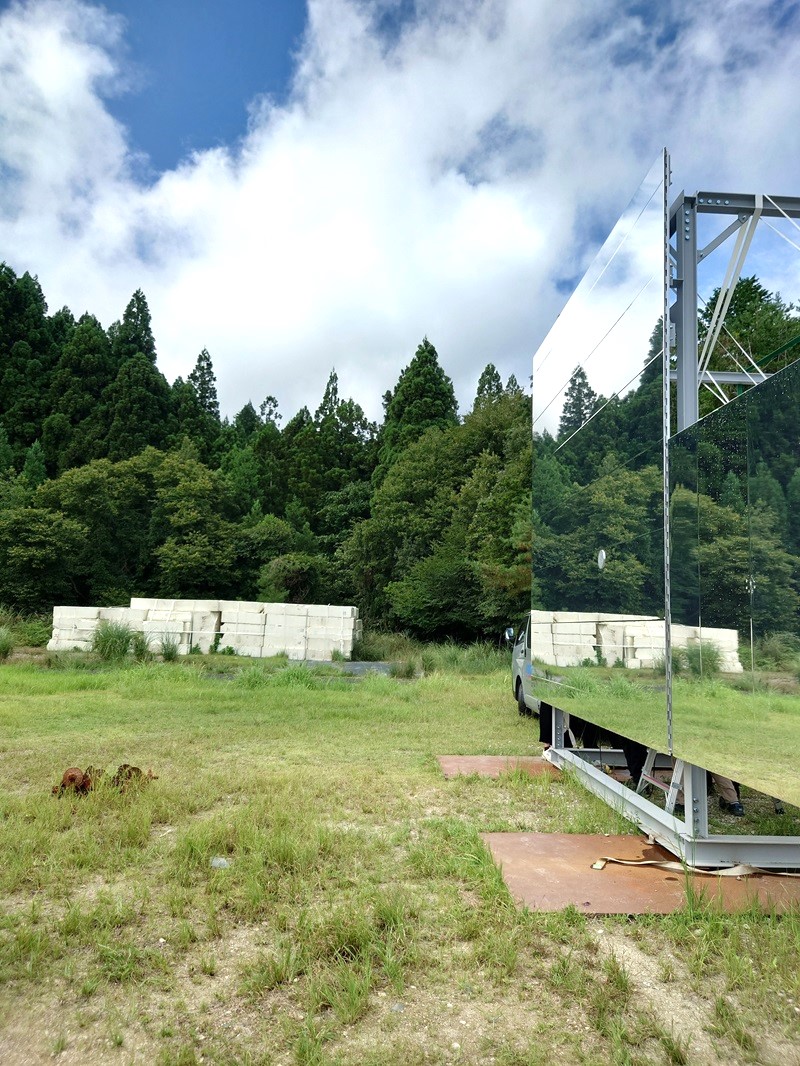 平面型は鏡そのものの出来栄えで、表面が揺らいでいないときは周囲の風景がそのまま映り込む。モックアップを設置した屋外の景色が映り込み、森もまた現実と映り込みの境目が曖昧に感じられる。万博会場では大阪・夢洲（ゆめしま）の空や周囲のパビリオンなどが映り込むことになる（写真：日経クロステック）